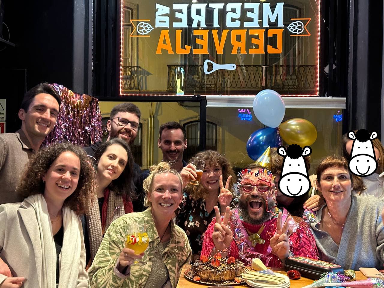 Pascal Pixel smiling and wearing a gold hat and comical birthday glasses, sitting at a table with a birthday cake and surrounded by a group of friends
