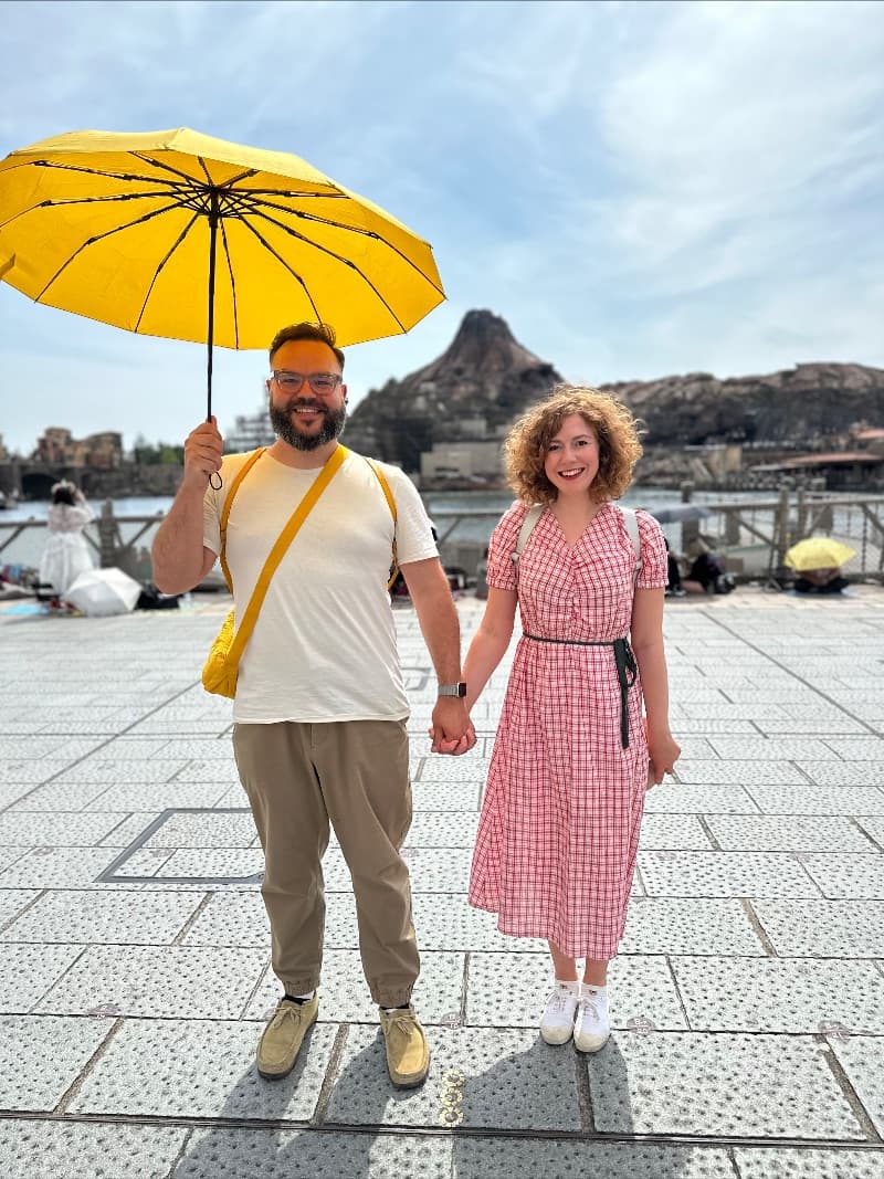 Pascal Pixel and Elly McKeown smiling and holding hands standing in front of a volcano attraction at Disney Sea in Tokyo, Japan